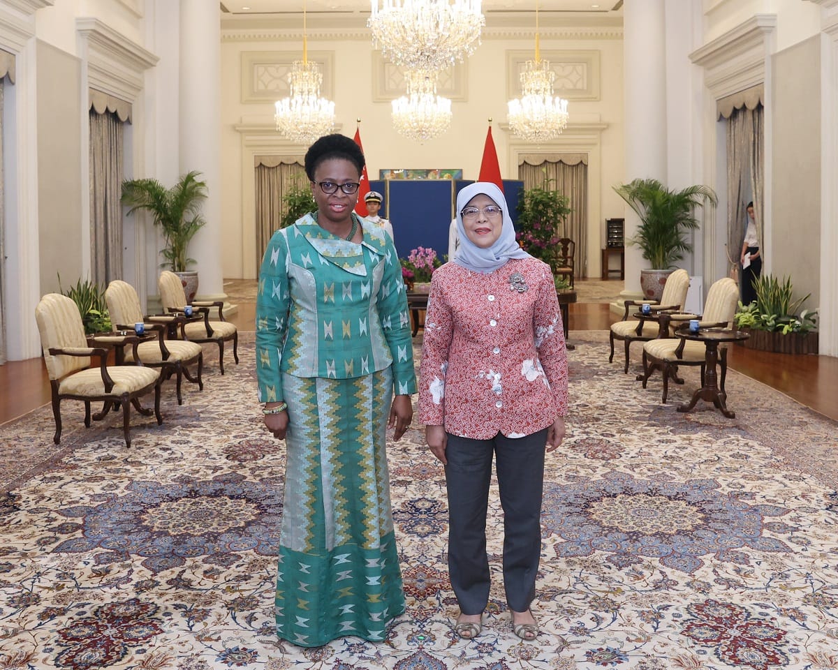 Two women stand on an ornate carpet in an ornate room with columns and chandeliers.